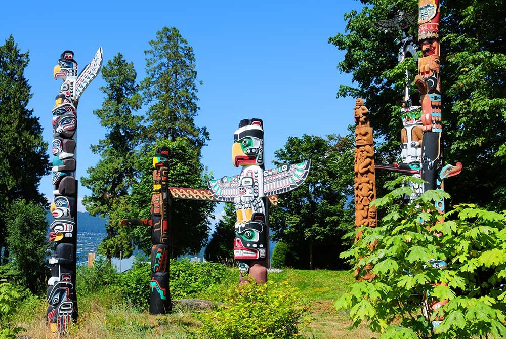 Four carved totem poles stand in a park with green trees and a clear blue sky, a highlight when you explore Vancouver attractions.